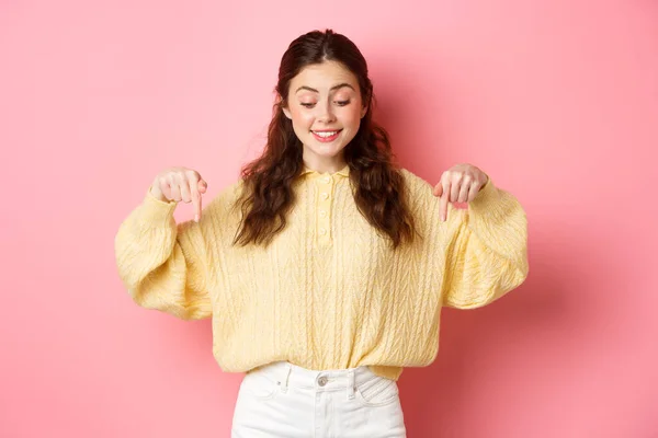 Joven mujer alegre en suéter, señalando y mirando hacia abajo con sonrisa feliz, intrigado por la oferta promocional, de pie sobre fondo rosa —  Fotos de Stock