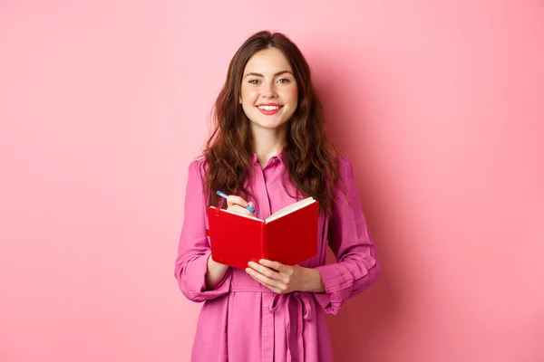 Fröhliches Mädchen lächelt und schreibt Notizen, hält ihren Planer, Plan oder Einkaufsliste in der Hand, steht mit Tagebuch vor rosa Hintergrund — Stockfoto