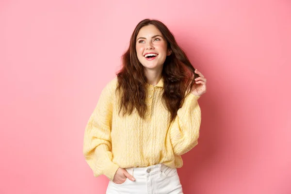 Imagem da jovem mulher dos anos 20 rindo e brincando com o cabelo, olhando para a promoção canto superior esquerdo, de pé feliz contra fundo rosa — Fotografia de Stock