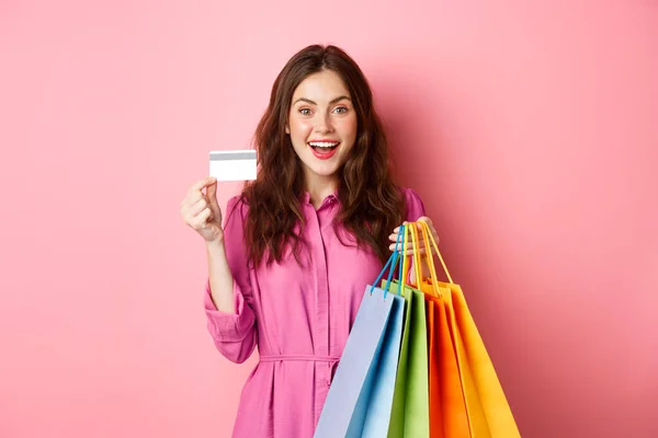 Porträt einer aufgeregten, fröhlichen Shopaholic, Frau mit Einkaufstüten und Plastikkreditkarte, erstaunt lächelnd, vor rosa Hintergrund stehend — Stockfoto