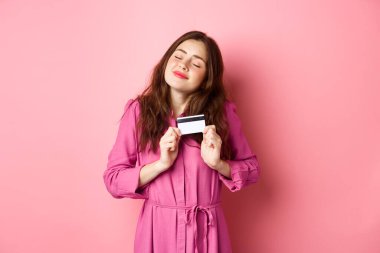 Genç esmer kadın, kredi kartını zevkle kucaklıyor, gülümsüyor, mutlu, pembe arka planda duruyor.