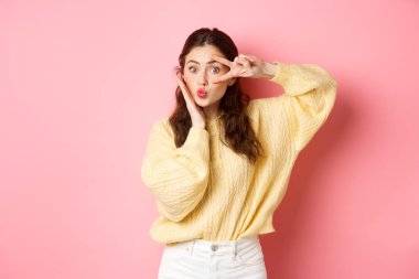 Tatlı aptal kız, öpüşürken yüzünü gösteriyor ve gözlerinin üzerinde V işareti var. Mükemmel yüz derisine dokunuyor. Pembe arka plana şaşırmış bir şekilde duruyor.