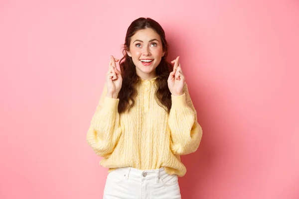 Симпатичная студентка желает сдать экзамен, скрестить пальцы, глядя вверх и улыбаясь оптимистично, стоя на розовом фоне — стоковое фото