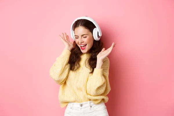 Chica feliz cantando canción favorita, escuchando música en auriculares inalámbricos, sonriendo y bailando, de pie sobre fondo rosa — Foto de Stock