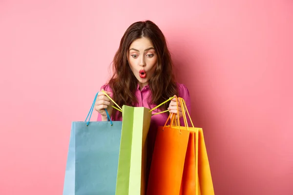 Aufgeregtes Mädchen öffnet Einkaufstüten und keucht erstaunt, schaut sich mit glücklichem Gesicht Geschenke für sie an, steht vor rosa Hintergrund — Stockfoto