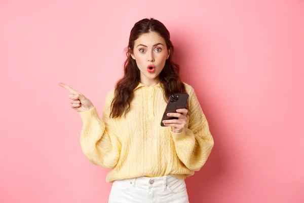 Förvånad ung vacker flicka kippar och säger wow, pekar finger åt vänster copyspace, håller mobiltelefon, står mot rosa bakgrund — Stockfoto