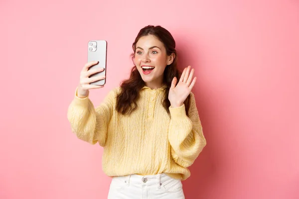 Веселая девушка видео чата на смартфоне, размахивая рукой перед камерой телефона и улыбаясь счастливым, сказать привет, стоя на розовом фоне — стоковое фото