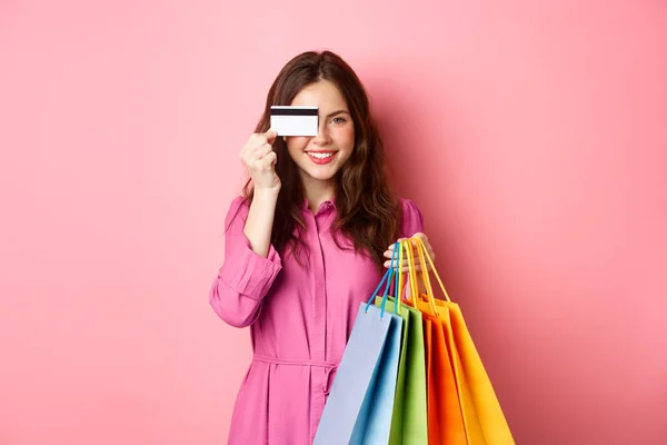 Portrét vzrušeného veselého shopaholika, žena držící nákupní tašky a ukazující plastovou kreditní kartu, usmívající se ohromeně, stojící na růžovém pozadí — Stock fotografie