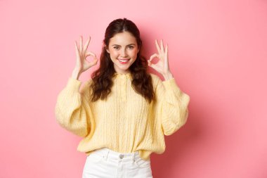 Kendine güvenen genç bir kız iyi işaretler gösteriyor ve gülümsüyor, mükemmel bir şey övüyor, evet ya da güzel bir jest diyor, seçimini onaylıyor, pembe arka planda duruyor.