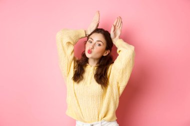 Paskalya tavşanı ya da tavşan kulağı gösteren tatlı ve aptal bir kız. Dudakları büzüşmüş, öpüşen surat yapıyor. Pembe arka planda duruyor.