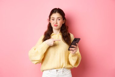 Şüpheci genç kadın tatminsiz suratını buruşturuyor, akıllı telefon ekranını işaret ediyor, online içerik hakkında şüpheleri var, pembe arka planda duruyor.