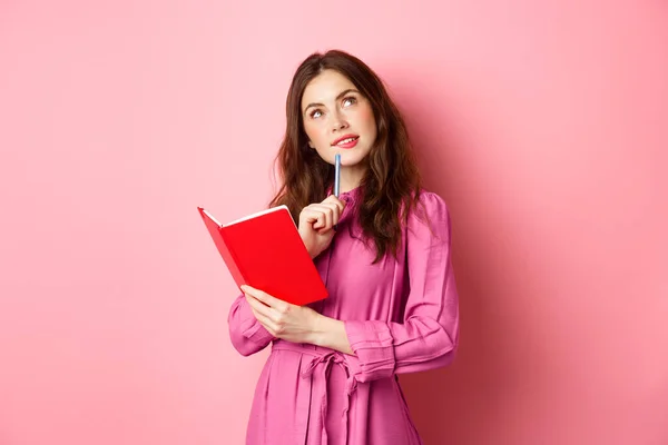 Schöne junge Frau sieht nachdenklich aus, schreibt in Notizbuch, hält Planer oder Tagebuch, plant ihren Zeitplan, steht vor rosa Hintergrund — Stockfoto