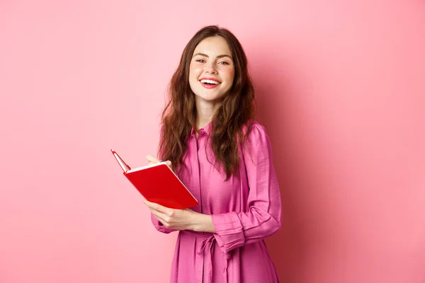 Beautiful young woman smiling, writing in notebook, holding planner or diary, standing against pink background — Stock Photo, Image