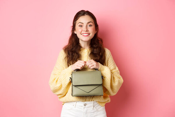 Beautiful lady holding her purse in hands near chest, smiling and looking excited at camera, waiting for something with temptation, standing against pink background