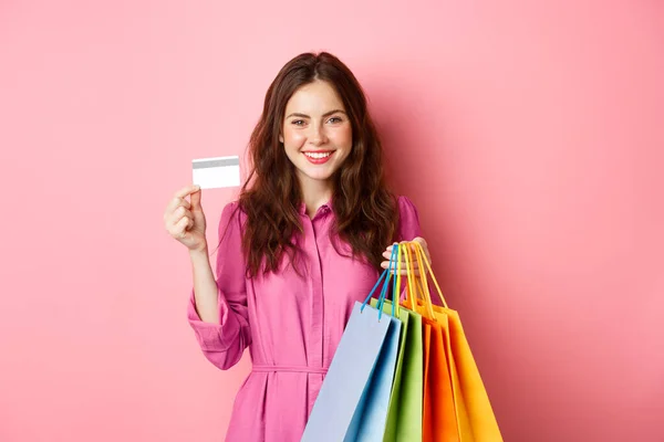 Junge brünette Frau hält Einkaufstüten, zeigt Plastikkreditkarte und lächelt, steht vor rosa Hintergrund — Stockfoto