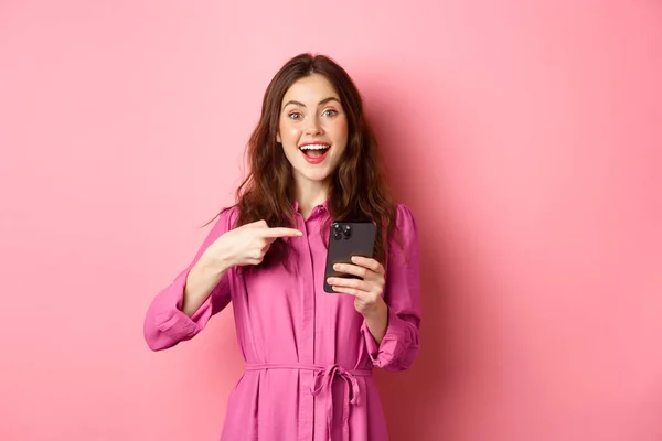 Young woman pointing finger at her phone and smiling, talking about smartphone app, showing something interesting online, standing over pink background — Stock Photo, Image