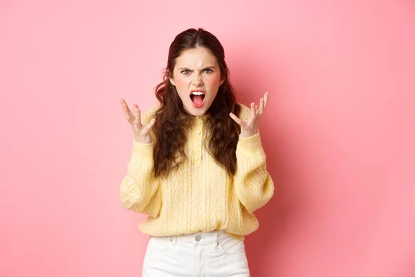 Angry and pissed-off girl screaming, shaking hands and grimacing furious, stare with outraged annoyed face at camera, standing against pink background — Foto Stock