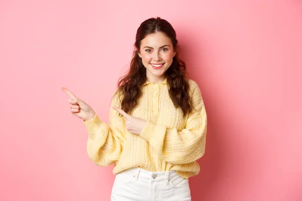 Retrato de mulher confiante sorrindo apontando dedos deixados no logotipo, mostrando texto promocional com determinado rosto, de pé contra fundo rosa — Fotografia de Stock