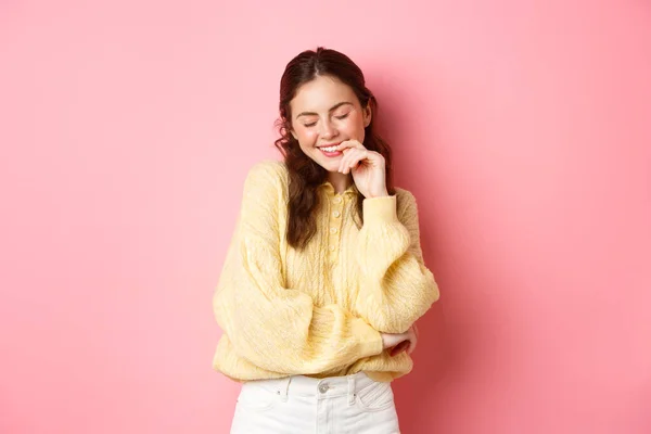 Nette junge Frau schließt die Augen und lächelt, errötet vor Kompliment, sieht verträumt aus, steht vor rosa Hintergrund — Stockfoto