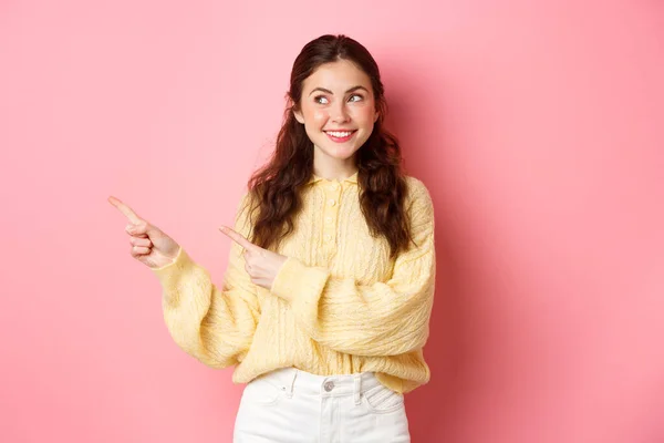 Молодая счастливая женщина улыбается, указывая и глядя в сторону на левый копирайт, показывая рекламу, стоя на розовом весеннем фоне — стоковое фото