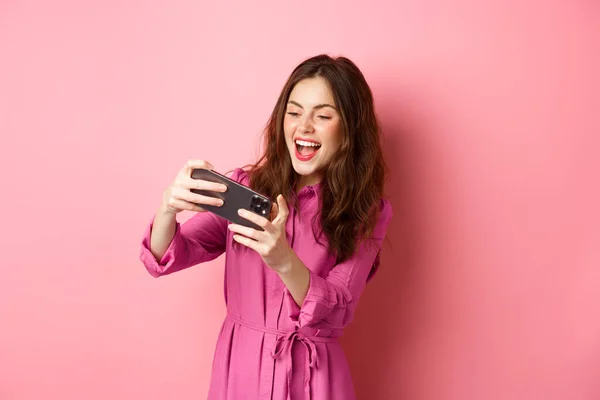 Технологическая концепция. Беззаботная молодая женщина наклоняет тело, держа смартфон горизонтально, играя в видеоигру по телефону, стоя на розовом фоне — стоковое фото