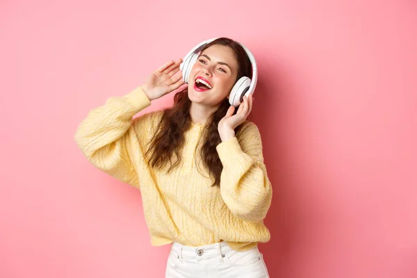 Hermosa chica moderna cantando canción favorita, escuchando música en auriculares inalámbricos, sonriendo y bailando, de pie sobre fondo rosa — Foto de Stock