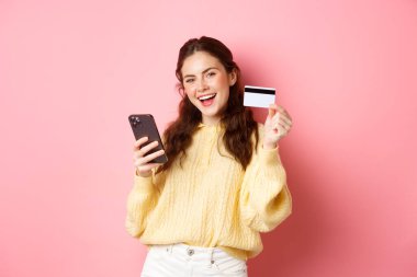 Teknoloji ve online alışveriş. Mutlu ve memnun bayan müşteri, plastik kredi kartı gösteriyor ve cep telefonu uygulamasını kullanarak online ödeme yapıyor, pembe arka plana karşı duruyor.