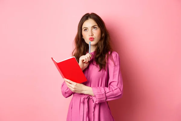 Beautiful young woman look thoughtful, writing in notebook, holding planner or diary, plan her schedule, standing against pink background — Stock Photo, Image