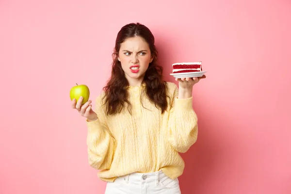 Menina saudável na dieta olha com nojo para pedaço de bolo, segurando verde aplicar, sorrindo insatisfeito, de pé contra o fundo rosa — Fotografia de Stock