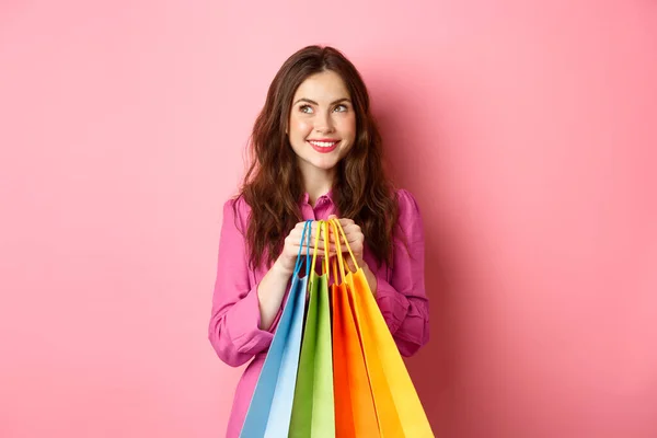 快乐的年轻女子想买东西，带着梦幻般的微笑抱着购物袋，买员工，站在粉红的背景上 — 图库照片