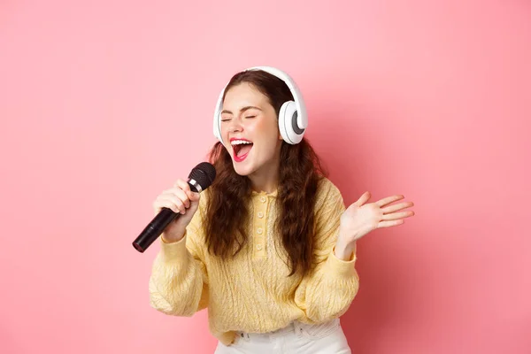 Retrato de una joven cantando karaoke, usando auriculares y cantando, sosteniendo micrófono, de pie sobre fondo rosa — Foto de Stock
