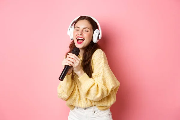 Chica despreocupada disfrutando de la noche de karaoke, cantando canción en el micrófono, usando auriculares inalámbricos, de pie sobre fondo rosa — Foto de Stock
