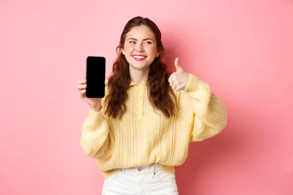 Bild av vacker kvinna skrattar och tittar åt sidan, samtidigt betygsätta en app, visar tummen upp och tom telefonskärm, rekommendera online promo, står mot rosa bakgrund — Stockfoto