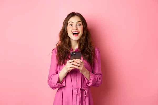 Beautiful brunette girl with amazed face, gasping and looking wondered, holding smartphone, see awesome news online, standing against pink background — Stock Photo, Image