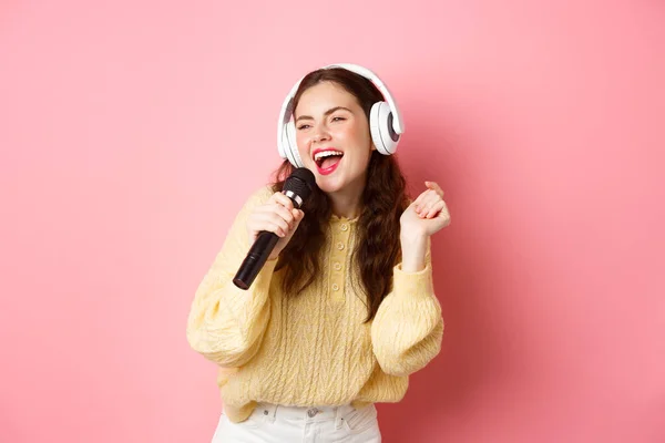 Ocio y estilo de vida. Chica morena feliz divirtiéndose, cantando karaoke en auriculares, sosteniendo el micrófono y mirando a un lado en la pantalla, de pie sobre el fondo rosa — Foto de Stock