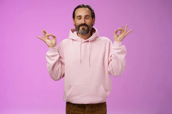 Friedliche charmante Hipster alter Mann trägt coole rosa Kapuzenpullover schließen Augen Atemübungen Stressabbau Meditieren im Stehen entspannt glücklich Yoga Meditation Pose Nirwana Geste, lila Hintergrund — Stockfoto