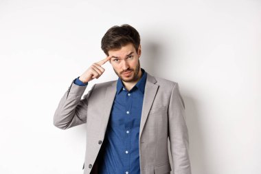 Sinirli erkek işverenin azarlaması, başını işaret etmesi, aklını mı kaçırdın sen? Alnının altından kameraya bakıyorsun, beyaz arka planda takım elbise içinde duruyorsun.