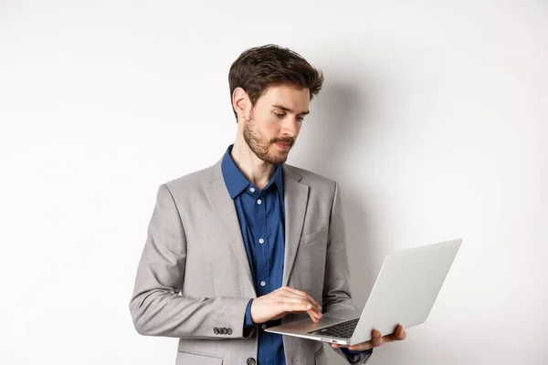 Knappe mannelijke ondernemer werken op laptop, op zoek serieus op het scherm, staan tegen een witte achtergrond — Stockfoto