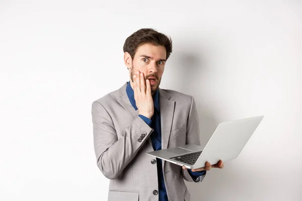 Geschokte en onrustige zakenman hebben problemen op de laptop, kijken bezorgd op de camera, maakte fout op het werk, staan op witte achtergrond in pak — Stockfoto