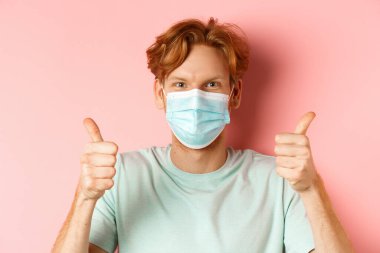 Covid-19 ve pandemik konsept. Dağınık kızıl saçlı yakışıklı, yüzüne tıbbi maske takmış, baş parmağını kaldırmış, pembe arka planda duruyor.