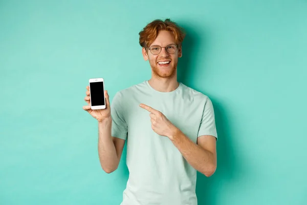Atractivo joven con barba roja y cabello apuntando con el dedo a la pantalla del teléfono inteligente en blanco, mostrando promoción o aplicación en línea, sonriendo a la cámara, fondo turquesa — Foto de Stock