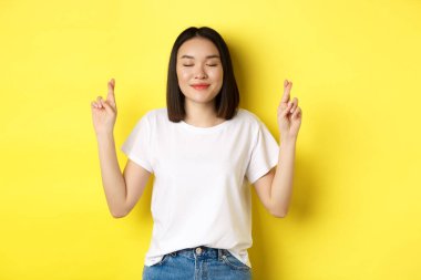 Umut dolu Asyalı kız, şans diliyor, şans diliyor ve gözleri kapalı dua ediyor, lütfen diyor, sarı arka planda duruyor.