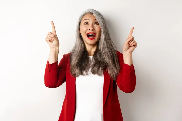 Glad asiatisk dam i röd blazer och makeup, tittar och pekar finger upp, visar specialerbjudande, står över vit bakgrund — Stockfoto