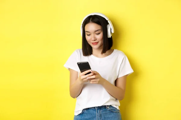 Fille asiatique moderne écoutant de la musique dans des écouteurs sans fil, lisant l'écran du smartphone et souriant, debout en t-shirt blanc sur fond jaune — Photo