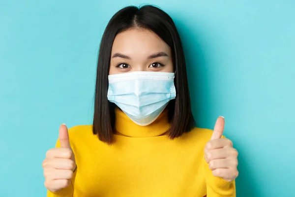 Covid-19, social distancing and pandemic concept. Close up of young asian woman in medical mask showing thumbs up, say yes, praise good offer, standing over blue background — Stock Photo, Image