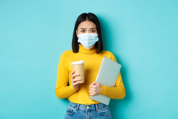 Covid-19, socialt avståndstagande och pandemikoncept. Orolig asiatisk kvinna i medicinsk mask, rynkar pannan ledsen, håller laptop för arbete och kopp kaffe — Stockfoto