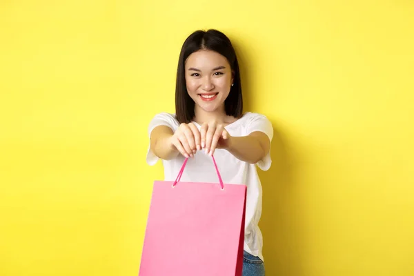 Nettes asiatisches Mädchen schenkt dir, streckt die Hand mit rosa Einkaufstasche aus und lächelt, gratuliert mit Urlaub, steht über gelbem Hintergrund — Stockfoto