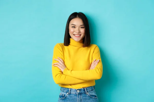 Kendine güvenen ve stil sahibi Asyalı kadın göğsünde çapraz kollar ve gülümsüyor, mavi arka planda duruyor. — Stok fotoğraf
