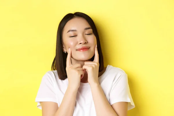 Beauty and skincare. Close up of young asian woman with short dark hair, healthy glowing skin, smiling and touching dimples on cheeks, standing over yellow background — Stock Photo, Image