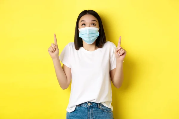 Covid-19, pandemiskt och socialt distanserande koncept. Ung asiatisk kvinna i vit t-shirt och medicinsk mask från coronavirus, tittar och pekar finger upp, visar specialerbjudande — Stockfoto